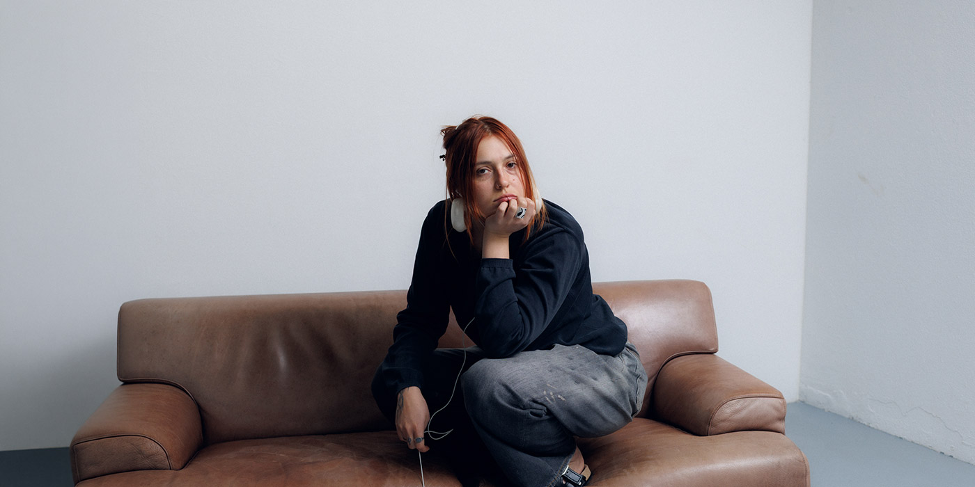 Die Sängerin LEILA hockt auf einer braunen Couch, die in einem leeren Raum steht.