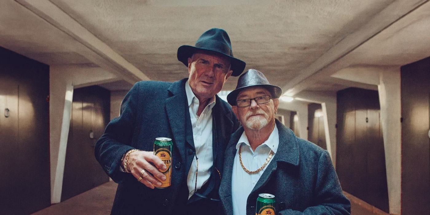 Pete und Bas, zwei Herren in ihren 70ern stehen Arm in Arm und mit Bierdosen in der Hand da. Sie tragen Hüte und Sakkos und versuche grimmig in die Kamera zu schauen-