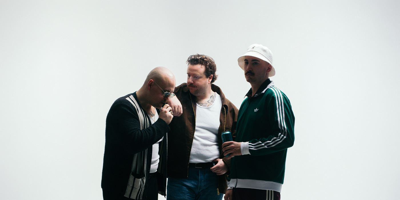 Die Glueboys, drei junge Männer sind zu sehen. Einer hat eine Glatze und Sonnebrille auf und zündet sich eine Zigarette an. Rechts daneben steht der zweite, und lehnt mit dem Arm auf dem ersten. Der dritte steht rechts davon in einem Adidas Jogger und weißem Kangol Hut mit eine Dose in der Hand.