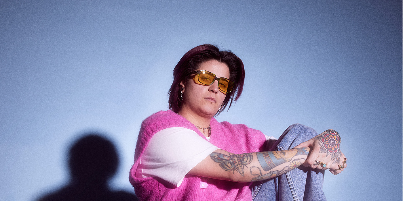 Schwesta Ebra sitzt auf einem Stuhl mit einem pinken Pulli und einer pink gefärbten Brille