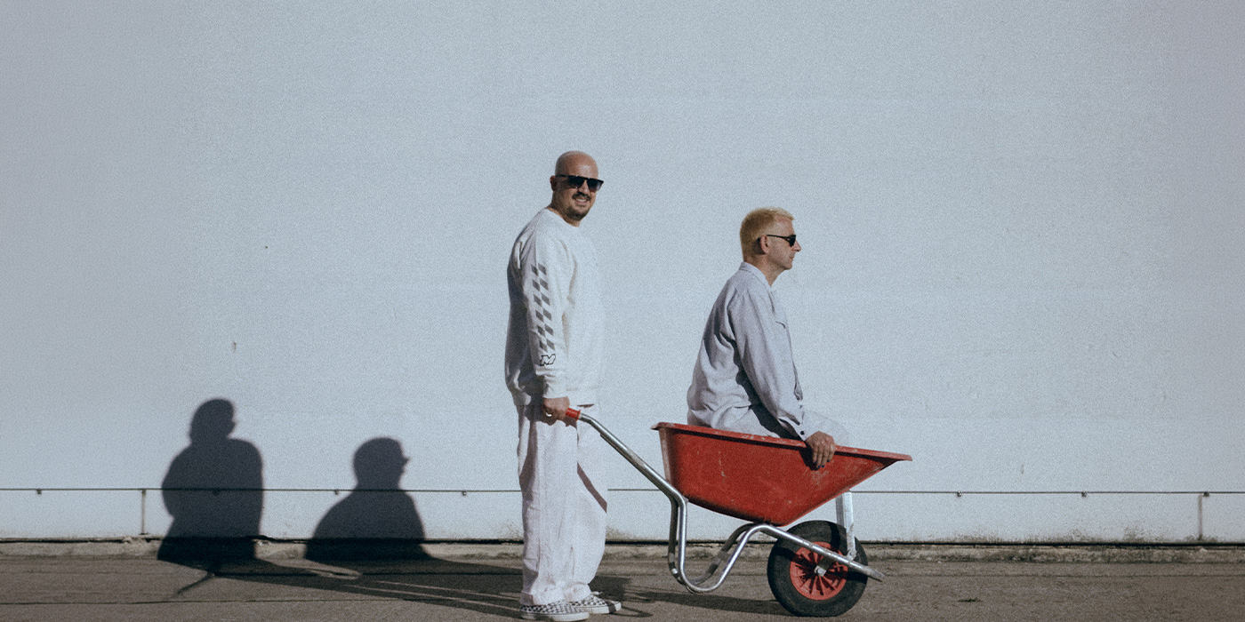 Das Duo von Modeselektor zeigt sich in dem einer in einer Schubkarre sitzt und der andre ihn schiebt. Beide sind in weiß gekleidet, vor einer weißen Wand, die Schubkarre ist rot.