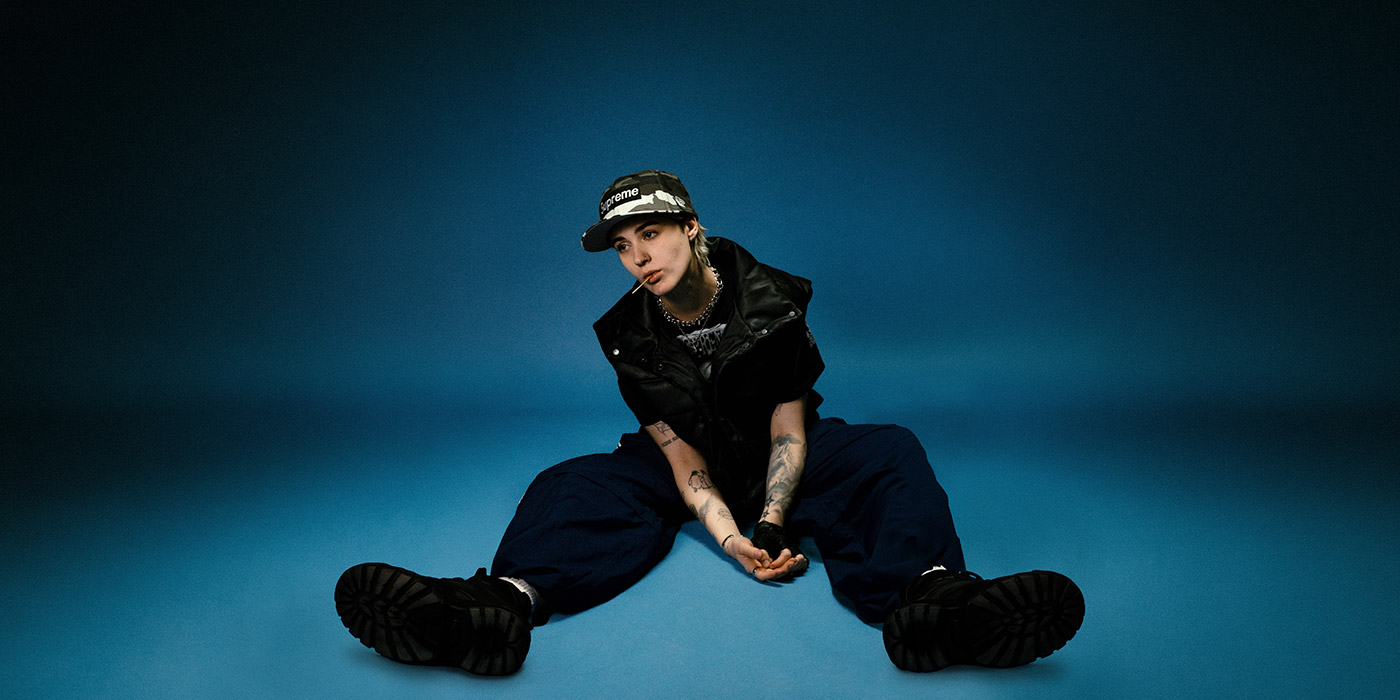 Artist NESS sitzt am Boden mit vor einem blauen Hintergrund