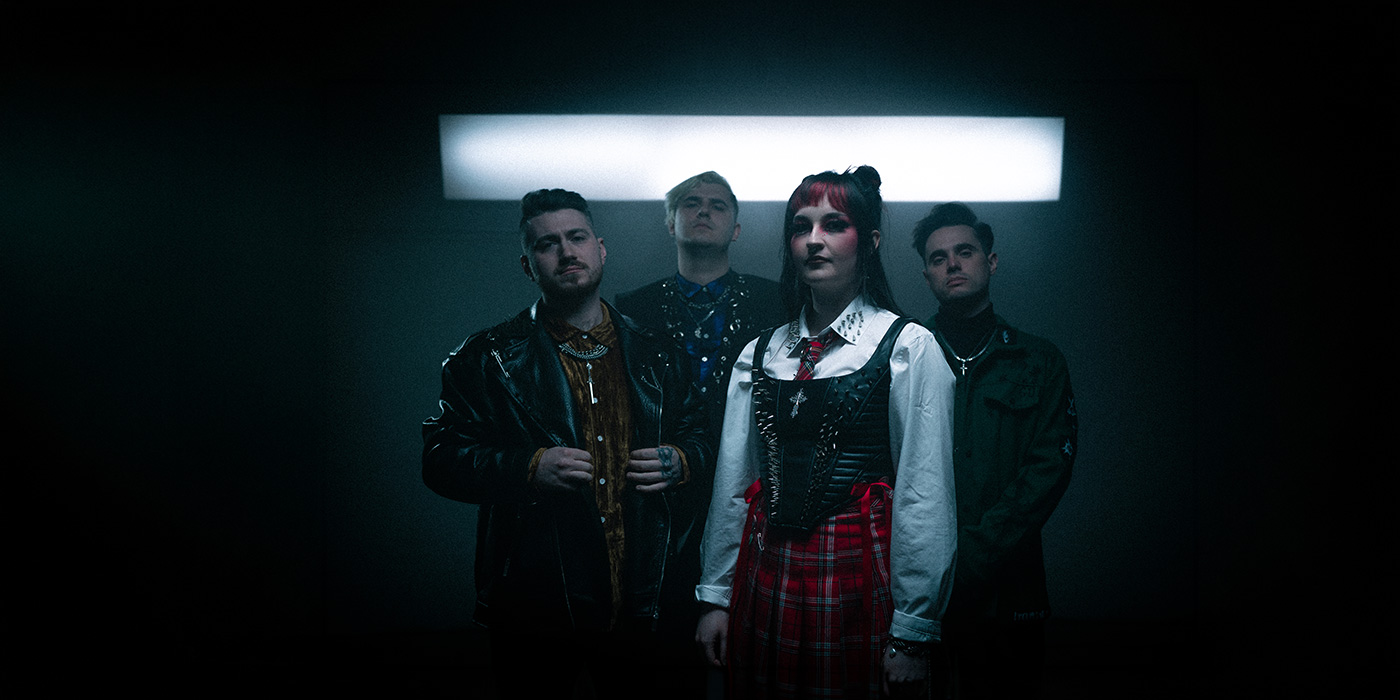 Die 4 Bandmitglieder von As Everything Unfolds schauen geradeaus in die Kamera. Dabei ist die Leadsängerin etwas heller beleuchtet in einer weißen Bluse, darüber ein schwarzes Korsett und eine Krawatte, gepaart mit einem Rock. Im Hintergrund sehen wir ein Licht.