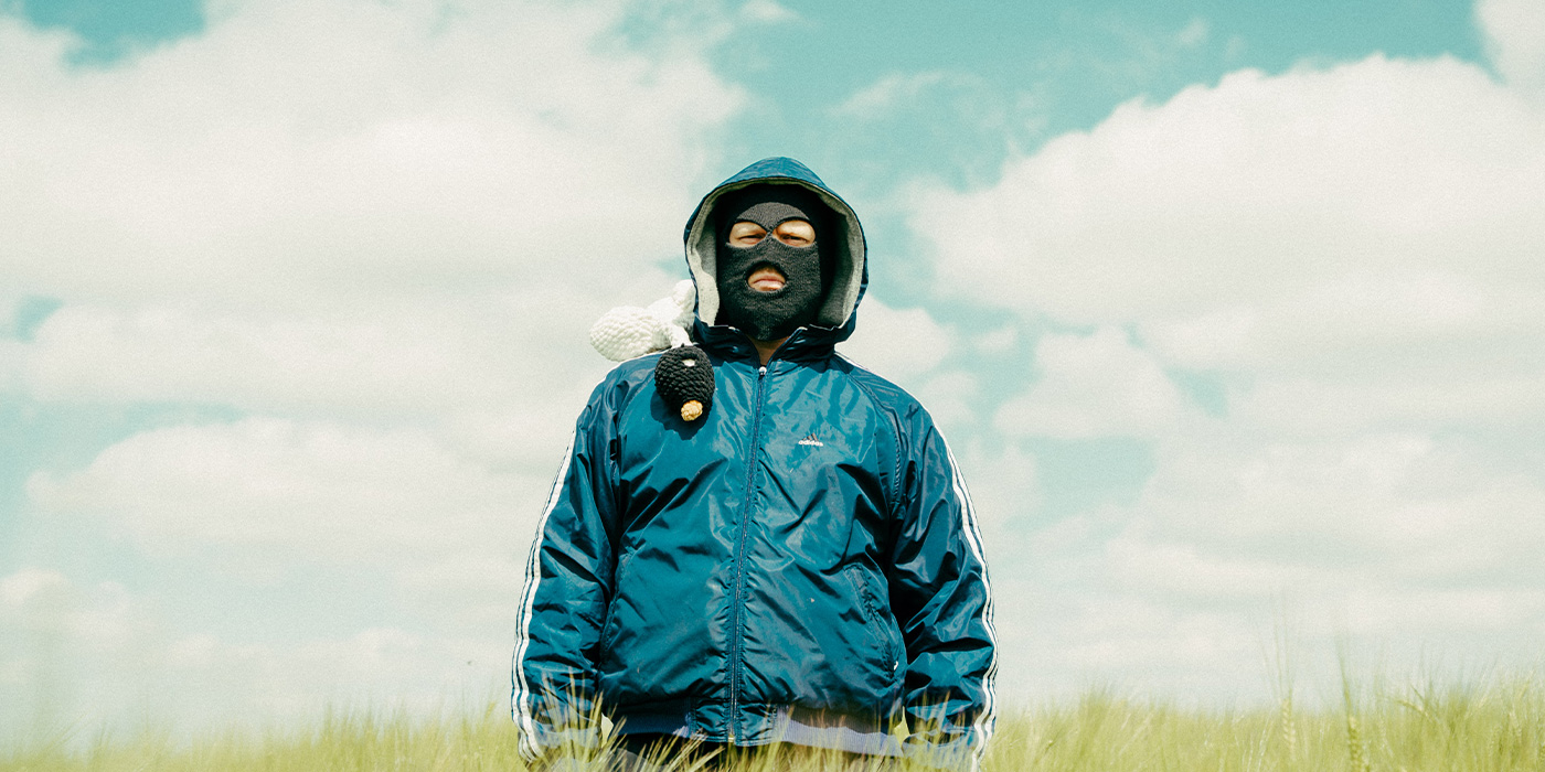 Der Künstler steht in einem Weizenfeld, das ihm bis zur Hüfte reicht. Sein Gesicht ist von einer schwarzen Sturmhaube zum Großteil unkenntlich gemacht. Er trägt eine petrolfarbene Adidas Jacke. Die Kapuze ist über den Kopf, über die Sturmhaube gezogen.