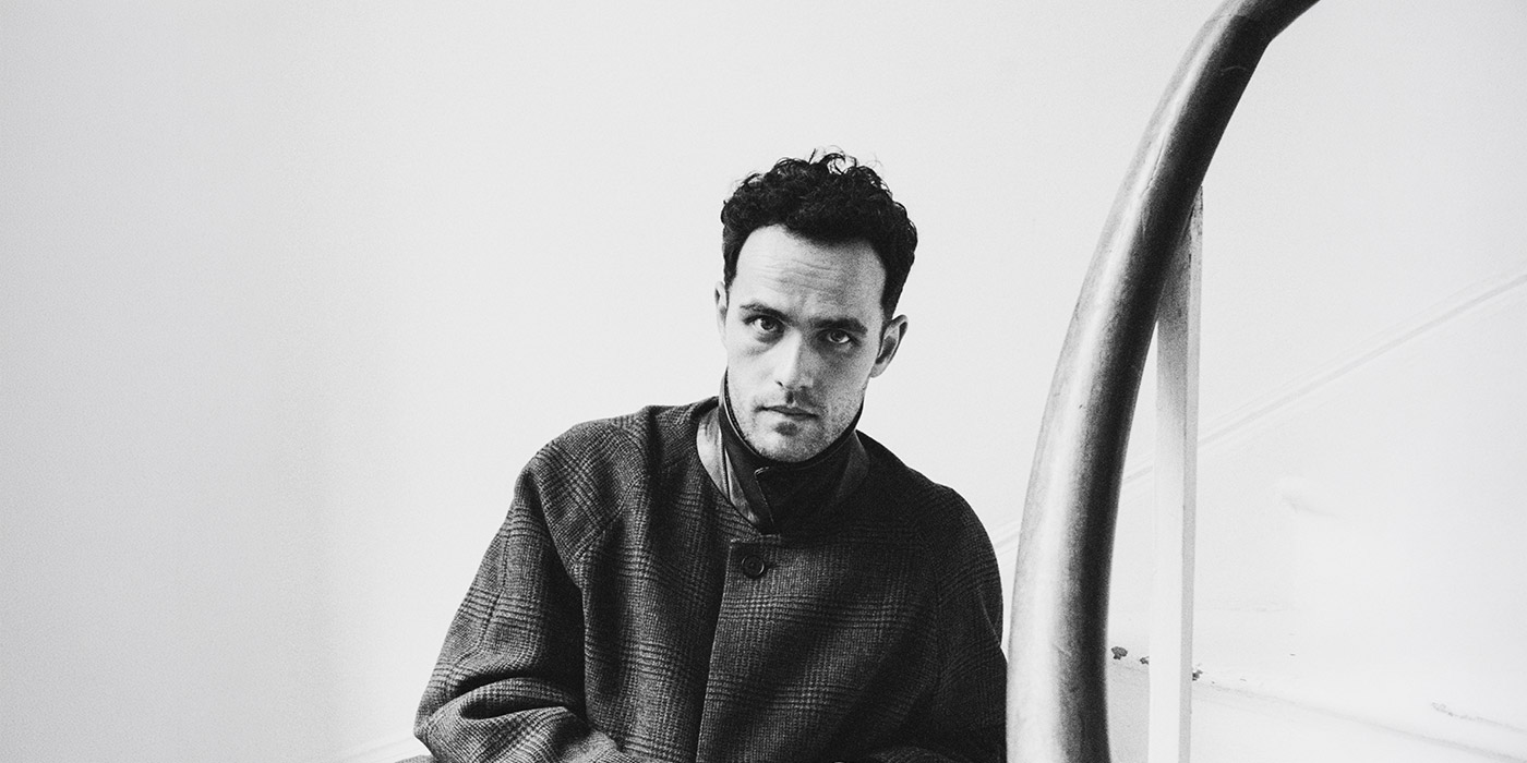 Jordan Rakei ist in schwarz-weiß zu sehen. Er blickt relativ ernst, mit leicht gerunzelter Stirn in die Kamera. Er trägt einen Tweed-Mantel.