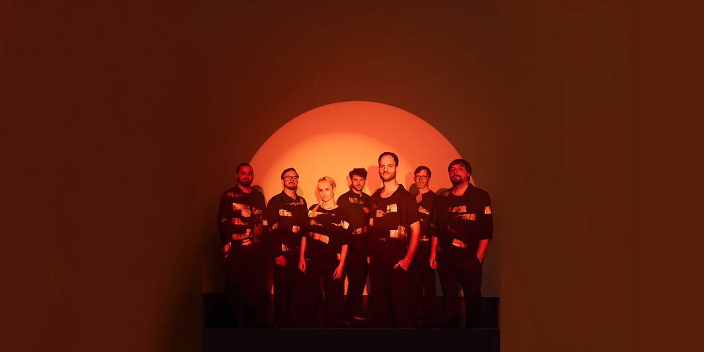 Die siebenköpfige Band wird mit einem einem Spotlight rötlich angeleuchtet. Über ihren Köpfen bildet das licht einen rötlichen Halbkreis, der an eine auf oder untergehende Sonne erinnert.