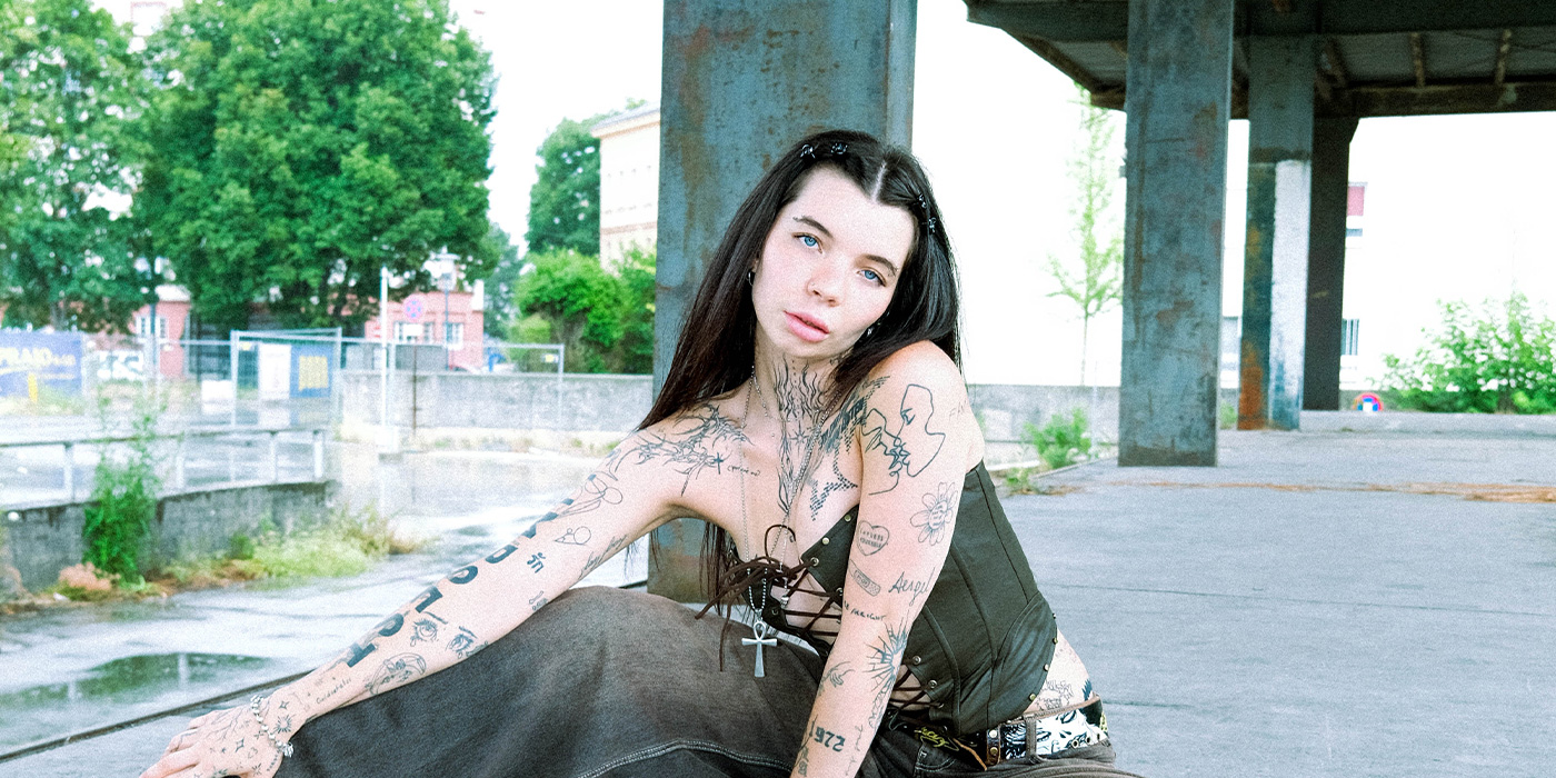 Luna Antonia sitz in urbaner Umgebung vor eine Betonsäule. Sie trägt Korsett, Jeans und ihr Oberkörper ist von vielen kleinen Tattoos übersät.