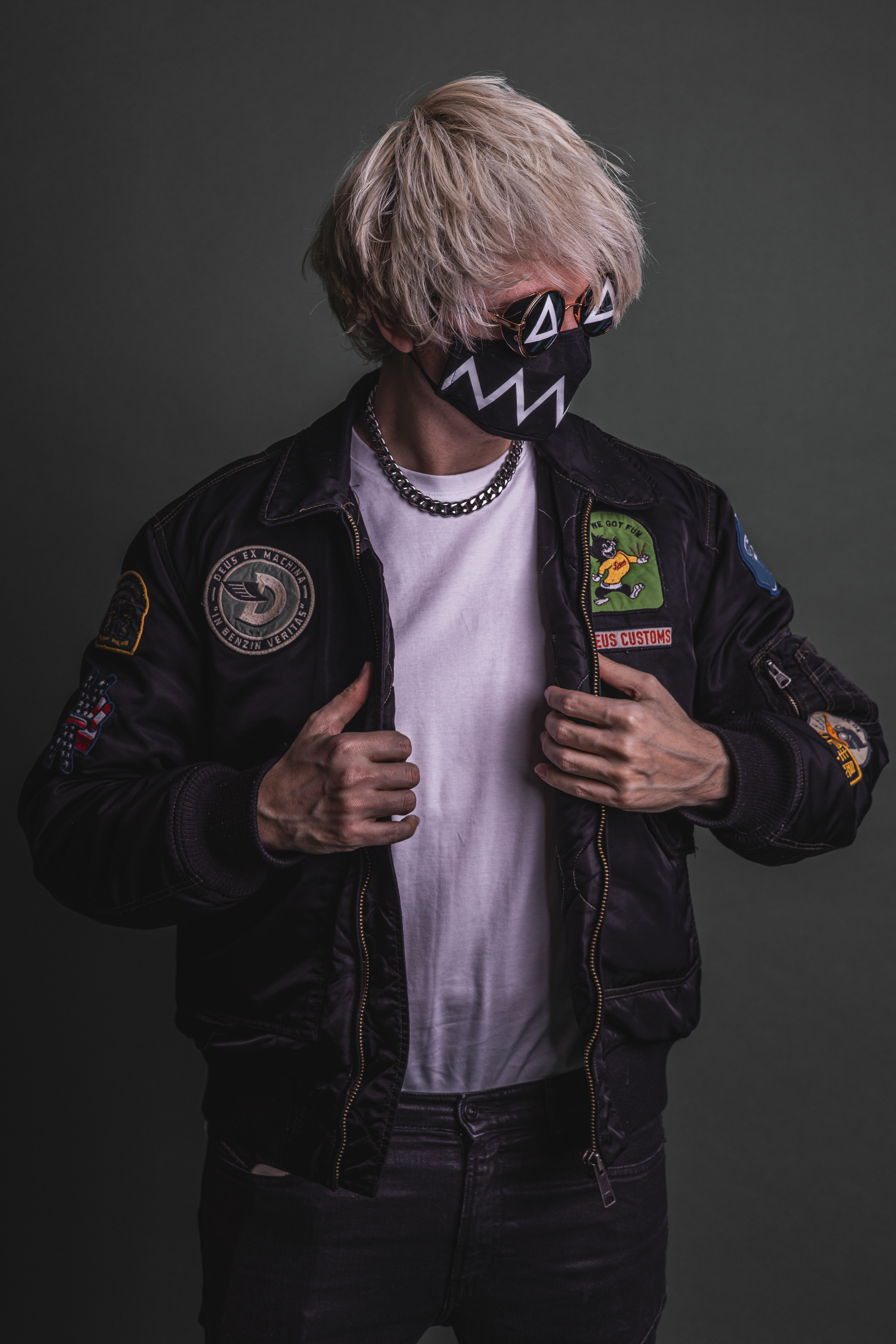 Tokyo Machine with his mask, looking away from the camera with his head. His body looking towards the camera, a white shirt, his arms grabs the dark jacket he wears.