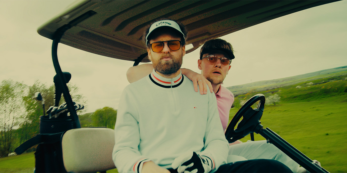 Die beiden Künstler sitzen in einem Golfwagerl, das sich auf einem Golfplatz befindet. Sie tragen weiße und rosa Golf Bekleidung und stylische Sonnenbrillen.