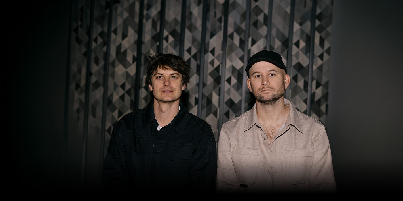 Camo&Krooked sind im Portrait vor einem gemusterten Hintergrund zu sehen.
