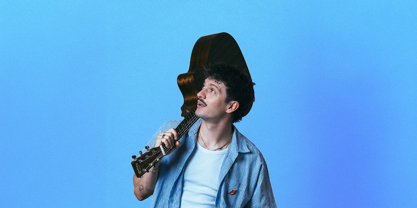 Der Künstler RIAN schaut in die aus zuschauerperspektive obere linke Ecke, über die Schulter hat er seine Gitarre gelegt. Er trägt ein blau gestreiftes Hemd, darunter ein weißes Shirt. Das Bild ist an den Oberarmen abgeschnitten, der Hintergrund in hellblau gehalten.