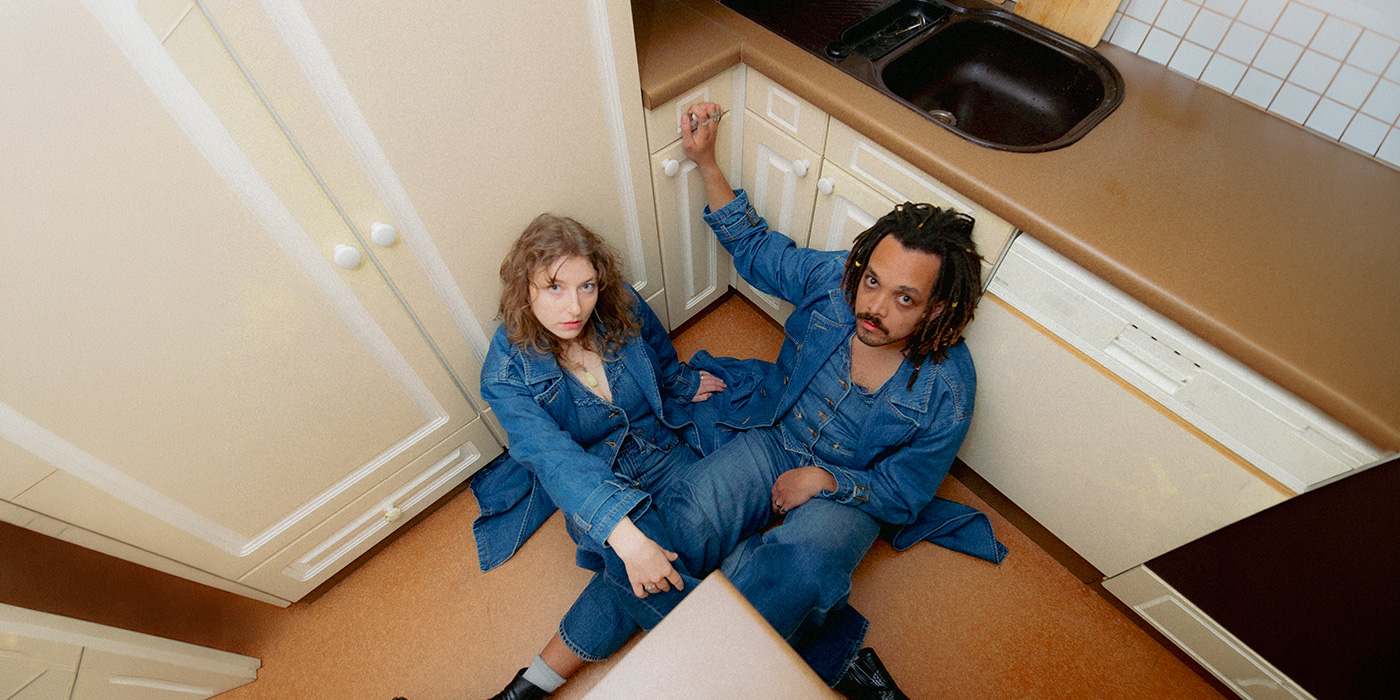 das duo sitz auf einem braunen Küchenboden. Sie tragen beiden Outfits aus Bluejean.