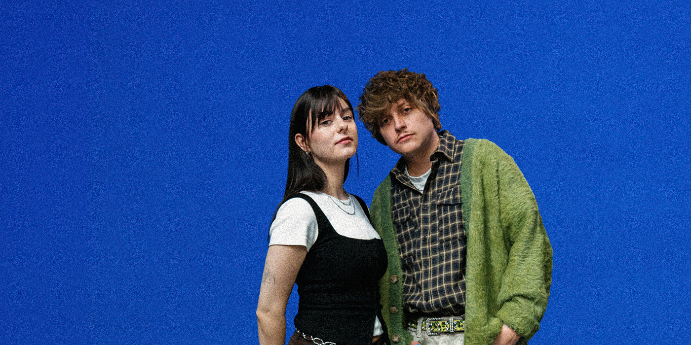 Die Artists Lena & Linus stehen vor einem knallblauen Hintergrund und schauen in die Kamera.