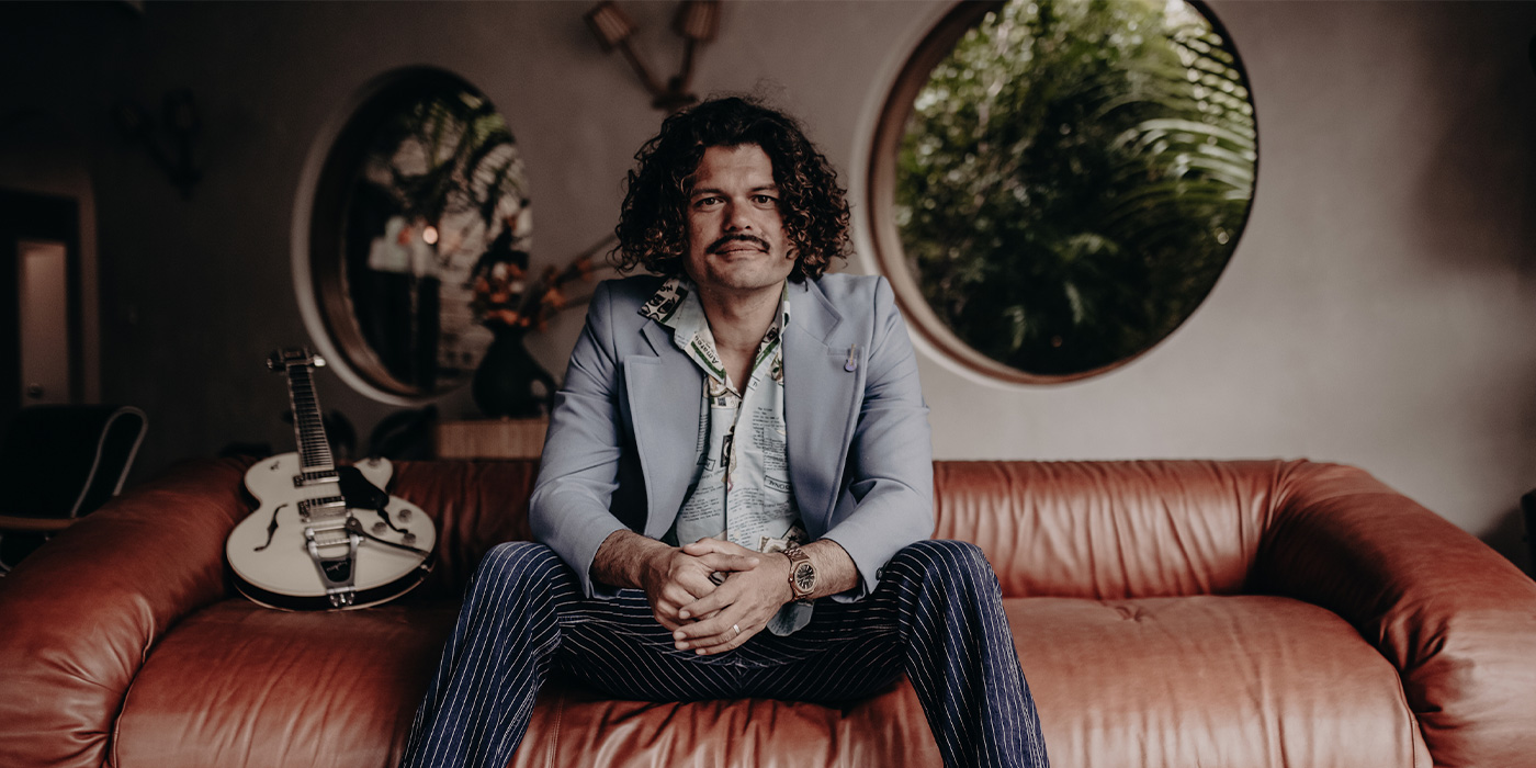 Ash Grunwald sitting on a brown leather coach, looking into the camera. Behind him are round windows looking into the green. Next to him is his guitar. While the background has a bit of a depth, we can see the artist clear. He has a dark blue, white striped pants, a shirt with patterns and a light blue blazer. His curly hair go until his shoulders and he has a sympathic smile on his face.
