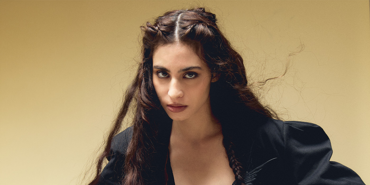 Tära is looking very intensively into the camera. Her brown hair are tied to braids. The background is light brown and in contrast to her dark hair and dark clothes this puts her into the foreground.