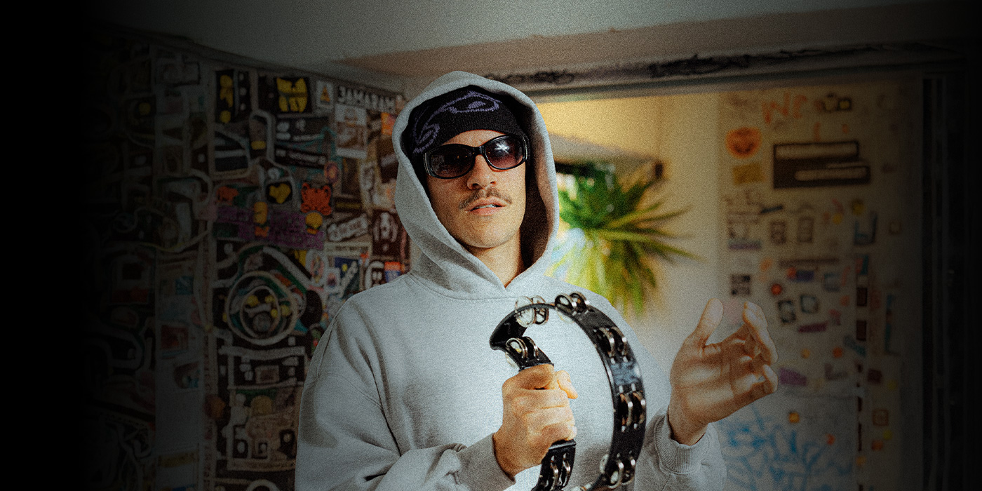 Der Artist ist vom Oberkörper aufwärts zu sehen. Er trägt einen grauen Hoodie, schwarze Sonnebrille und schwarze Haube unter der Kapuze. In den Händen hält er ein Tamburin