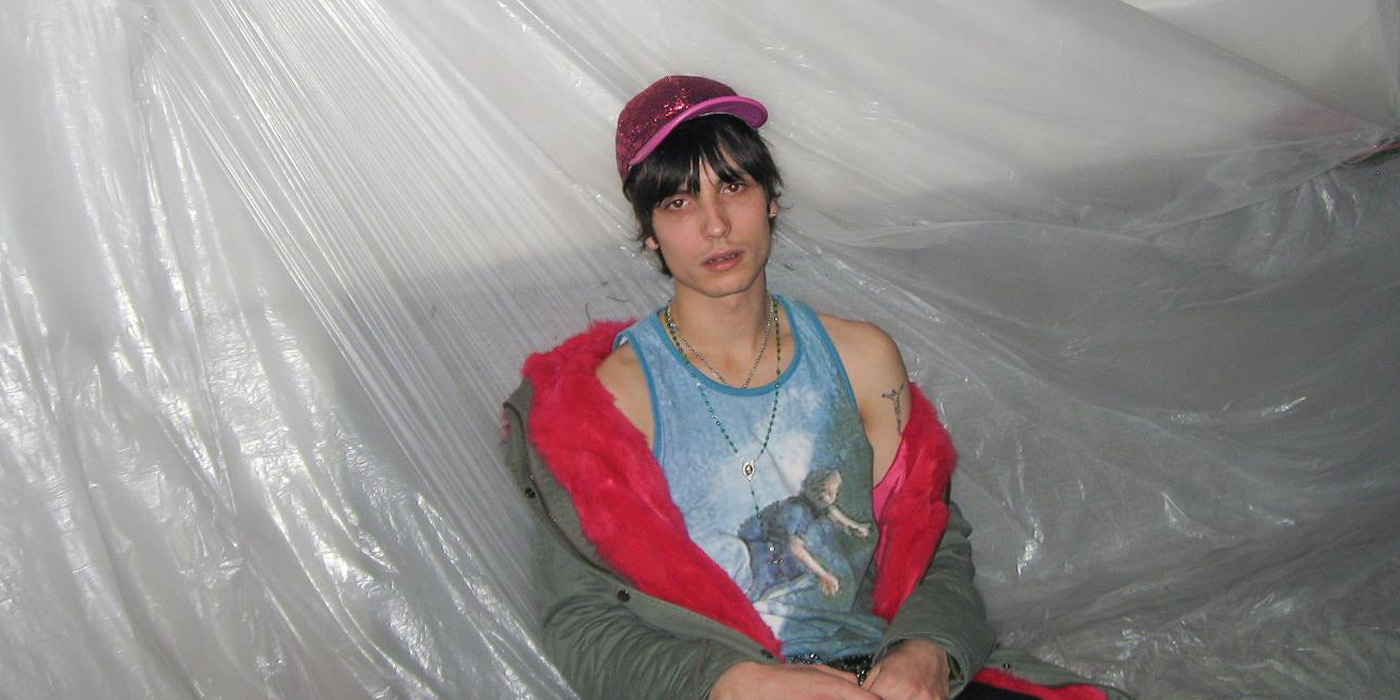 Der Künstler sitzt auf einer Plastikfolien-ähnlichen Oberfläche. Er trägt eine pink glitzernde Kappe, ein hellblaues Trägershirt und eine Jacke mit pinkem Innenfeld die ihm von den Schultern gerutscht ist.