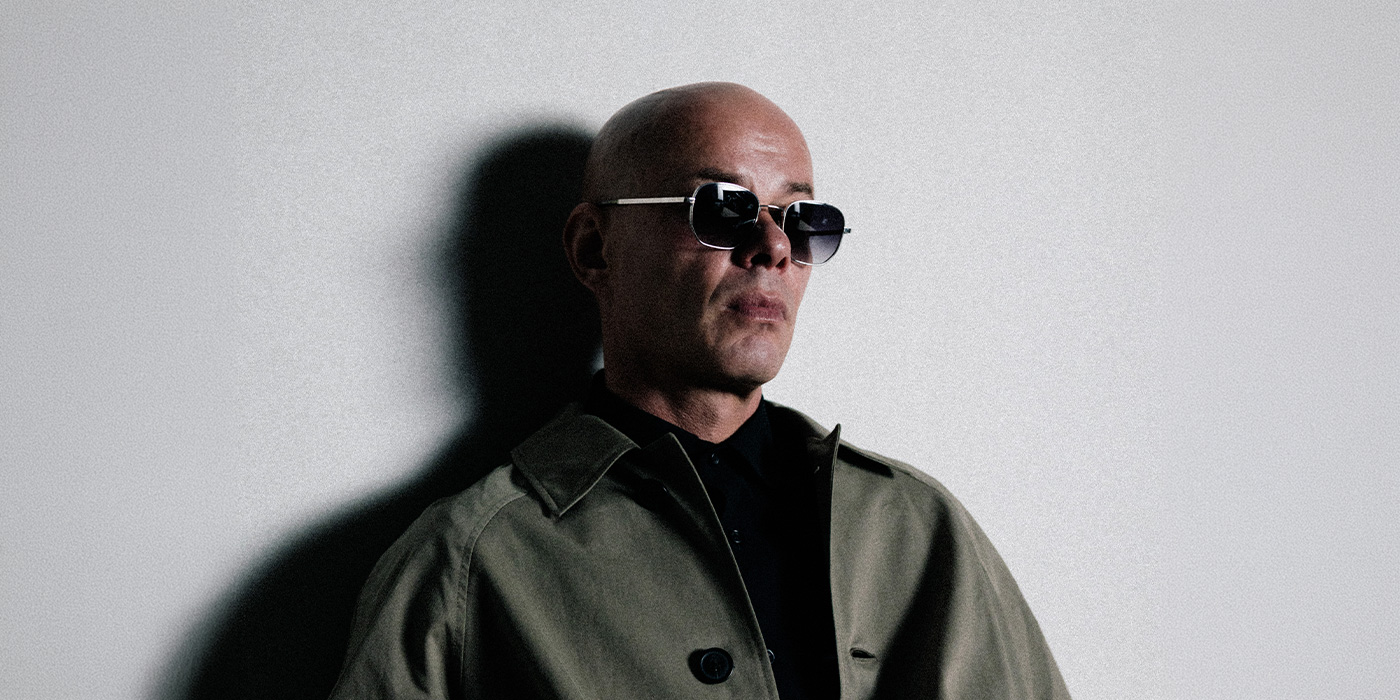 Trettmann trägt eine silber umrandete Sonnenbrille und olivgrünen Trenchcoat. Er lehnt ab euber weißen Wand, auf die sein Schatten fällt.