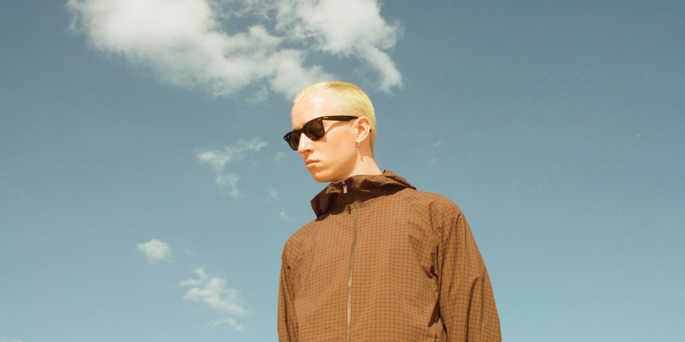 Jack Kays hat blondes Haar und eine Sonnenbrille auf. Er trägt eine braune Jacke. Der Hintergrund ist ein blauer Himmel mit vereinzelten Wolken.