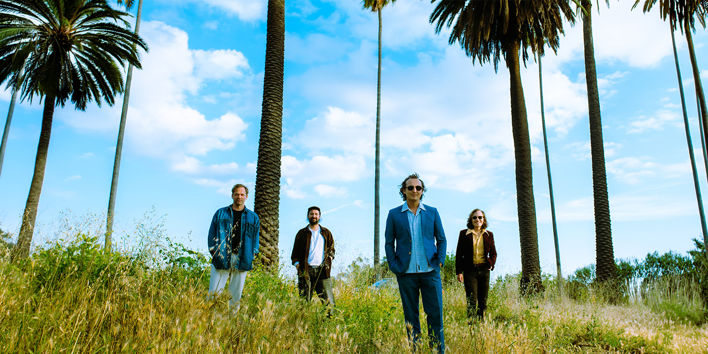 Die Band Theodor steht im Gras zwischen Palmen, jeweils immer versetzt. Der Himmel blau mit Wolken. Alle schauen glücklich in die Kamera