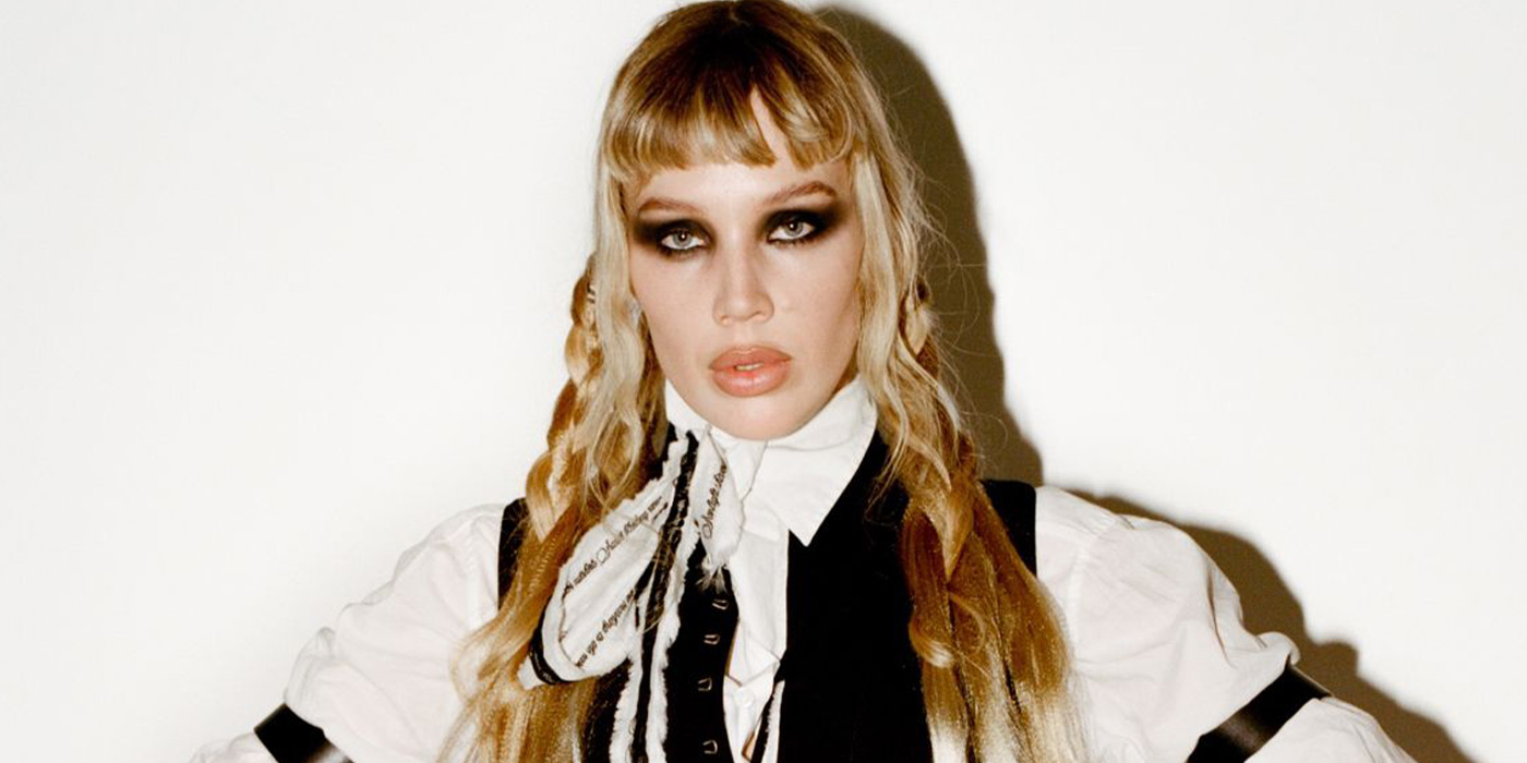 Die Künstlerin ist vor einem weißen Hintergrund abgelichtet. Sie trägt lange blonde Haare mit kurze Stirnfransen, eine Art schwarz-weiße Bluse und hat dunkel geschminkte Augen.