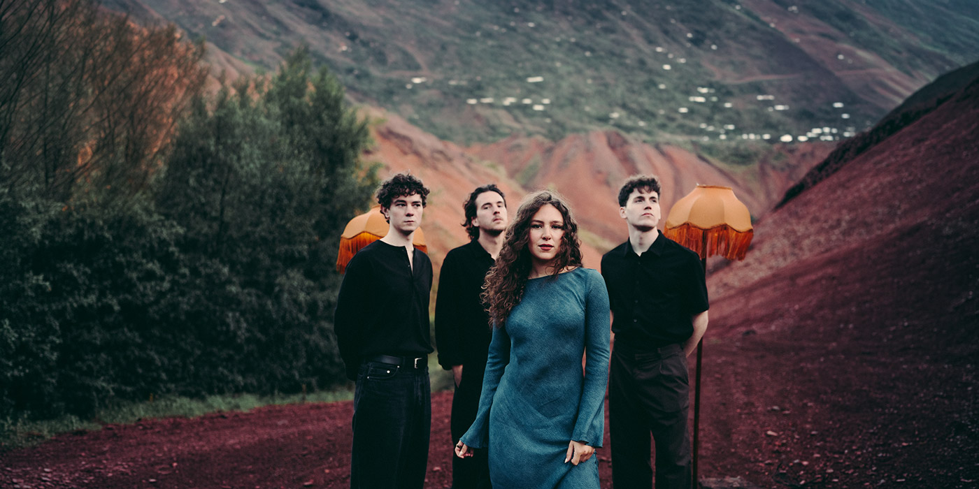 Fright Years posing in a red rocky surrounding, in the background they have set up lamps. The lead singer Jules Kelly is standing in the front with a blue dress, Fraser Charles (bassist), Harrison McLeod-Bonnar (guitarist) and Struan Blacklock (drummer) are wearing black and standing behind her.