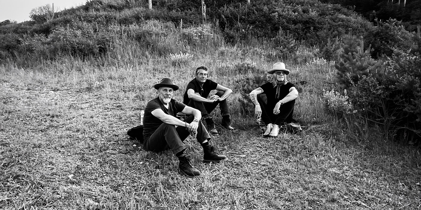 schwarz weiß foto der band wie sie auf einer wiese sitzt. beine aufgestellt, Arme auf den Knien abgelegt. Zwei der drei Mitglieder tragen Hüte in Westernstyle