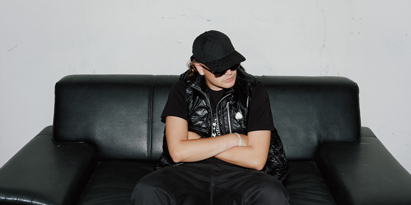 YungSaintPaul sitzt auf einer schwarzen Couch. Er hat eine schwarze Cap und schwarzes Shirt, drüber ein schwarzes Gilet. Mit einer Sonnenbrille auf der Nase schaut er in die rechte Ecke aus der betrachtenden Perspektive.