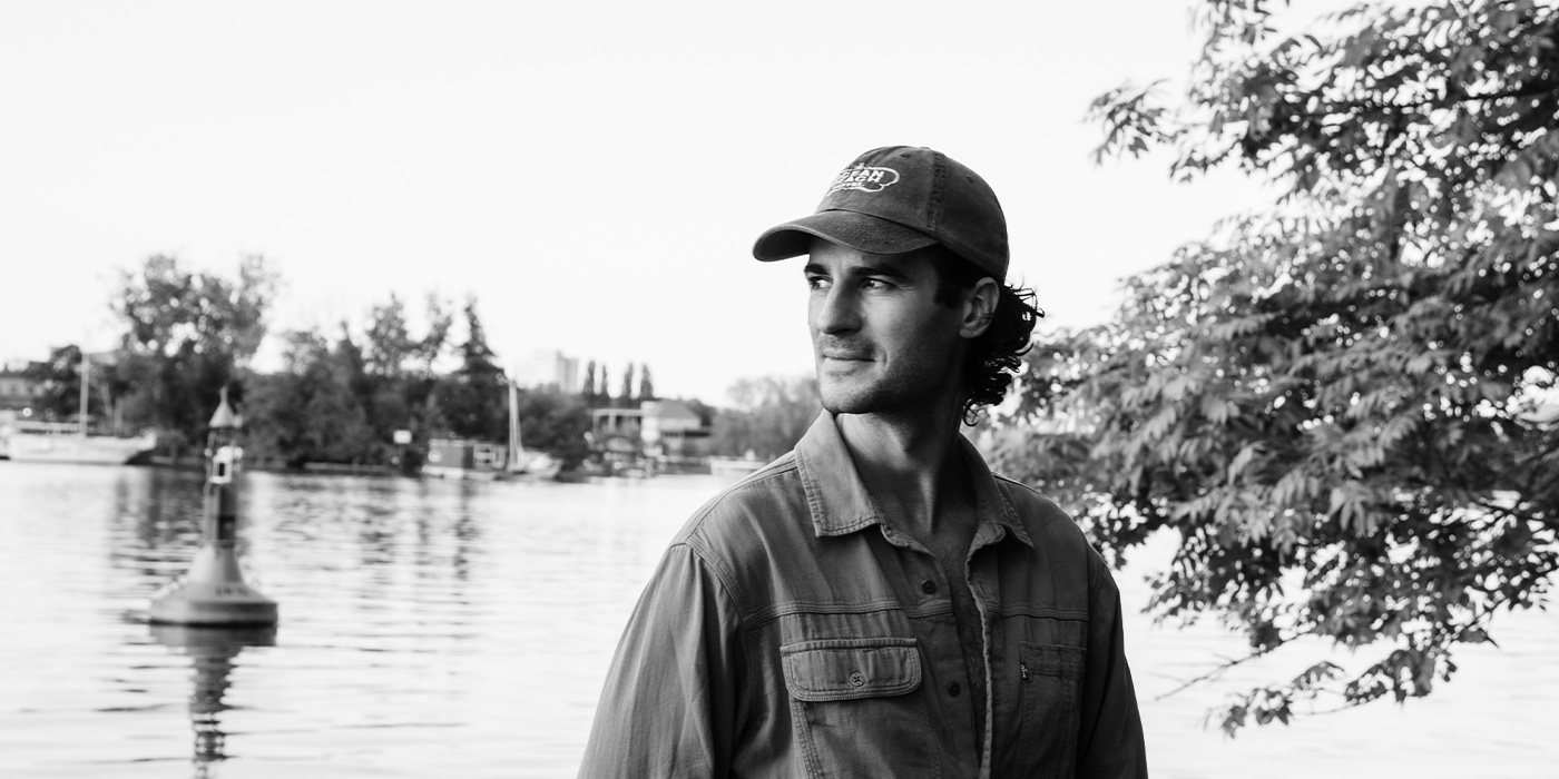 Ben Camden can be seen in a black and white photo. He is standing in front of a body of water, wearing a jeans shirt and a baseball cap. He is looking to the side.