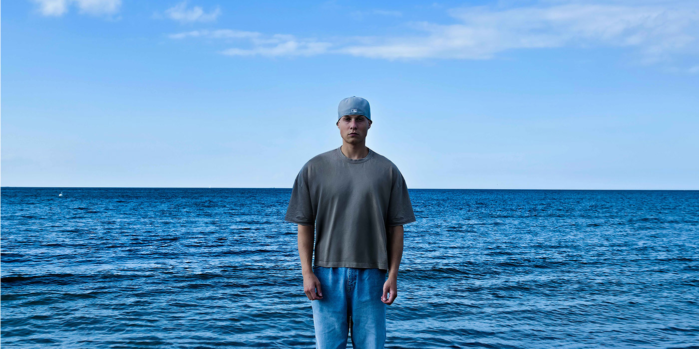 Matterz steht mit Kappe, grauem Shirt und blauer Jeans vor einem blauen Meer mit hellblauem Himmel. 