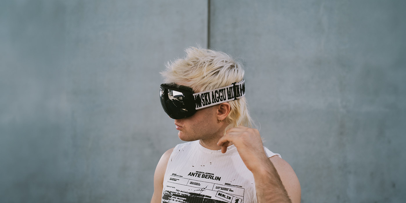 Ski Aggu ist, wie immer, mit blondierten Haaren und Skibrille tragend abgelichtet. Er blickt nach links und trägt ein schwarzweißes ärmelloses Shirt.