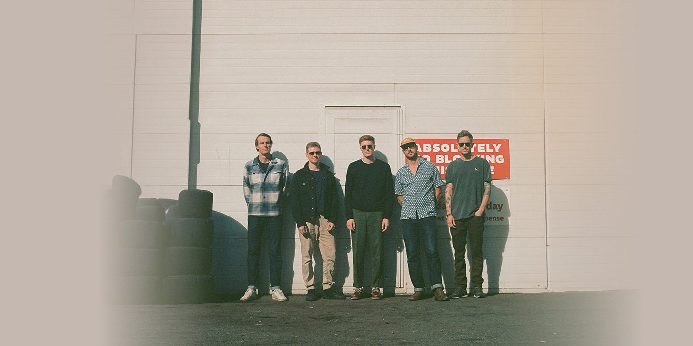 Band members can be seen standing in front of a white warehouse wall. to their left a bunch of stacked tires can be seen.