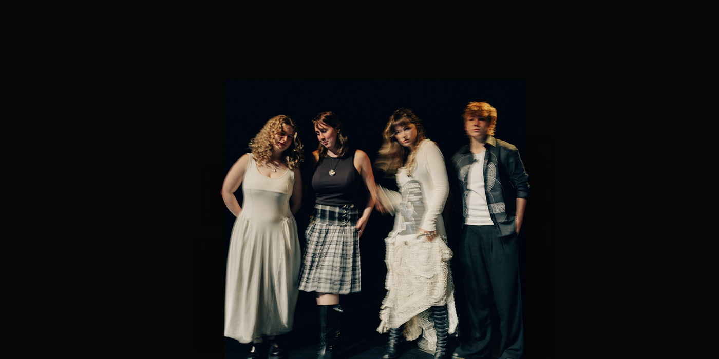 a blurry photo if the four-piece band. the background and floor are black, the band members are dresse im white and blue, mostly dresses and skirts.