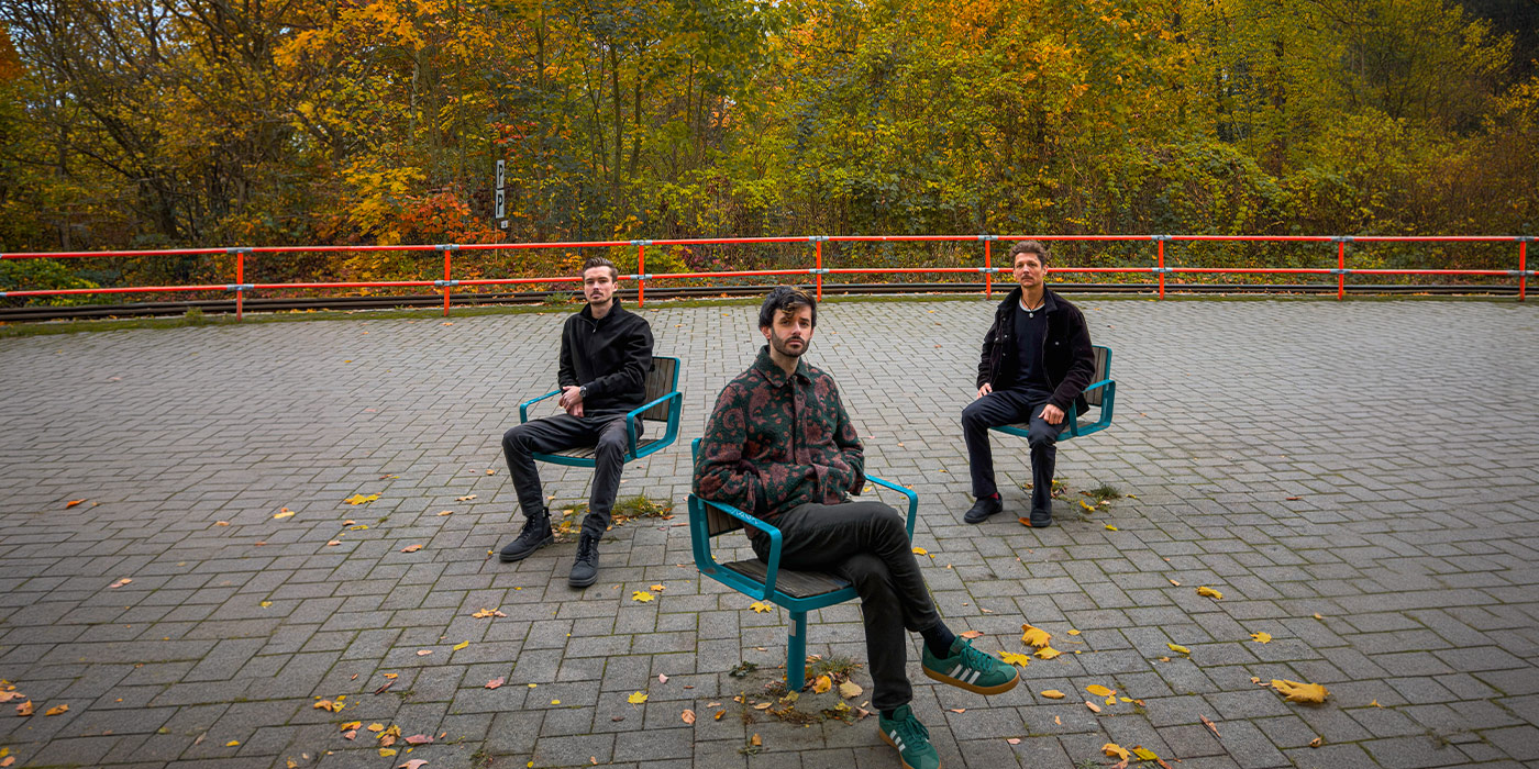 Three band members are sitting in a public space. Behind them colorful autumn leaves, a red railing in front of the trees and the band sitting on three single chairs.